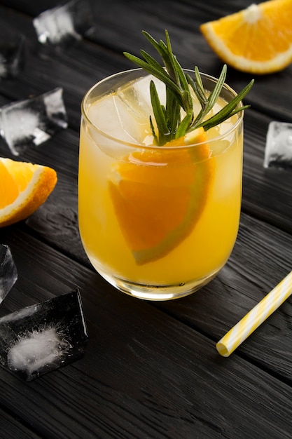 Orange drink or lemonade with rosemary and ice  in the glass  on the black wooden background. Closeup. Location vertical.