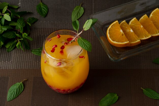 Orange drink with mint ice and pomegranate on a brown table with orange slices on a plate