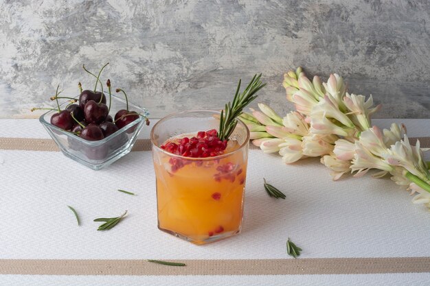 orange drink with red pomegranate and a branch of rosemary on a table accompanied by cherries