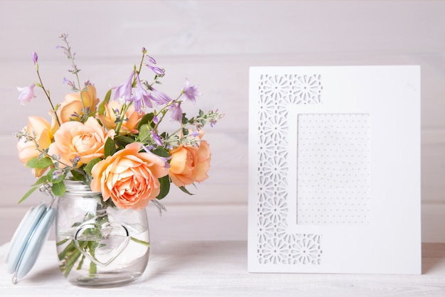 Orange English roses flowers in vase. jar and empty white photo frame on a wooden background. Mockup with copyspace.