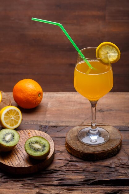 Orange fruit cocktail with a straw garnished with a slice of lemon in a large glass on a wooden table next to oranges and kiwi