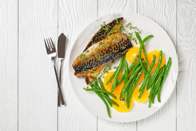 Orange glazed mackerel fillet with fresh orange and steamed green beans on a white plate with cutlery on a table flat lay free space