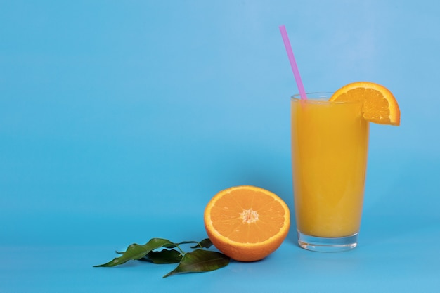 Orange juice in a glass with a straw, decorated with a slice of orange, half an orange with leaves on a blue background. Summer drink. Copy space