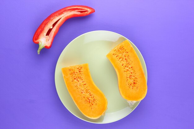 白いプレートにオレンジ色のジューシーなカボチャの半分と赤ピーマンの半分紫色の背景にカラフルな野菜のクローズ アップ 上からの眺め