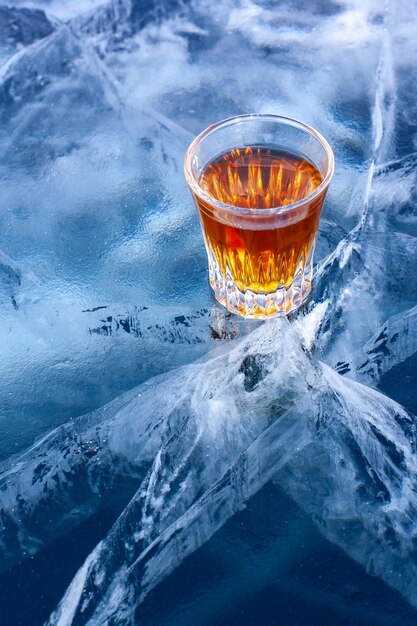 Orange liquid in a glass stands on the beautiful ice of Lake Baikal Cold whiskey in the winter Blue ice with beautiful deep white cracks Top view from the side Vertical