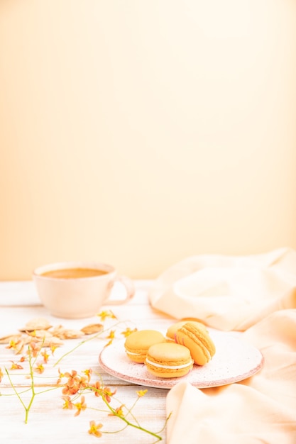 Orange macarons or macaroons cakes with cup of apricot juice on a white wooden background and orange linen textile.