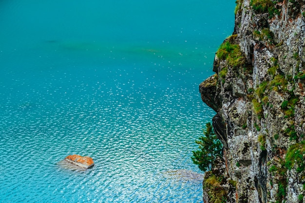 Photo orange mossy stone in azure mountain lake near large cliff with rich vegetation