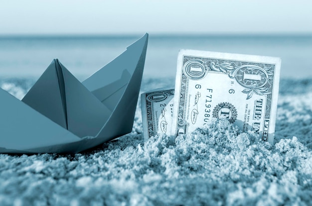 Orange paper boat and two paper dollar bills half buried in sand on sandy beach