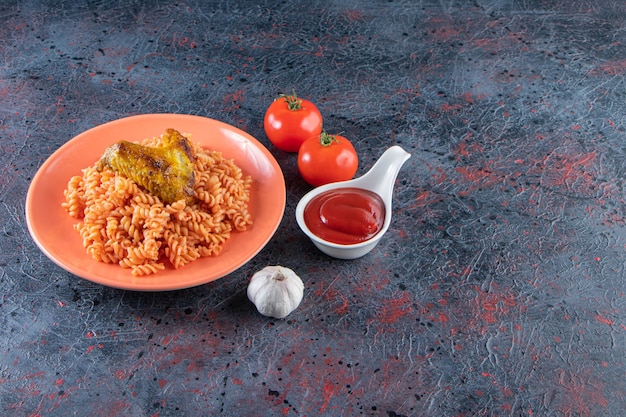 Orange plate of fusilli pasta with chicken wing on marble surface. 