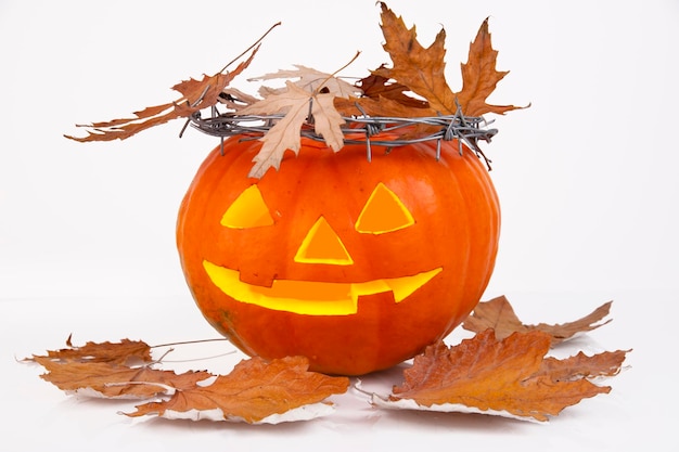 Orange pumpkin with glowing eyes nose and mouth with a crown of\
thorns barbed wire and dry maple leaves on a white background\
halloween