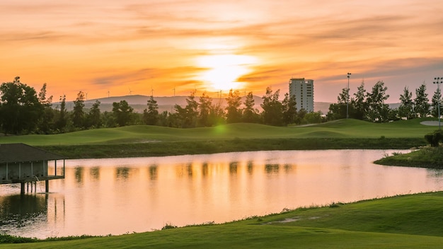 Orange red dawn sun above horizon closeup lake pond foreground water texture surface calm reflection trees high building background park city hills green grass mountains fabulously mysterious