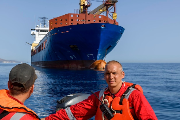 Foto imbarcazione di salvataggio arancione con equipaggio in mare con una grande nave da carico blu sullo sfondo trapano uomo a mare addestramento su scialuppe di salvataggio