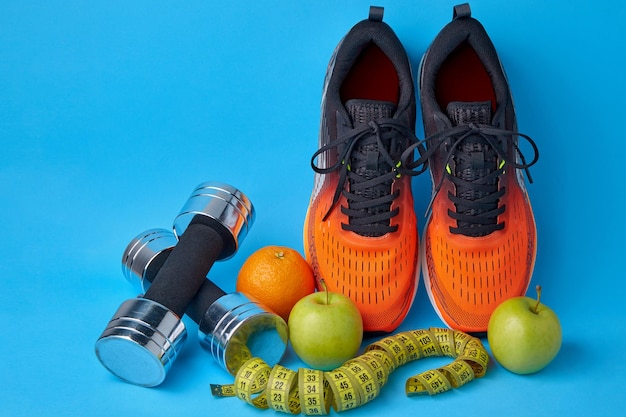 Orange sneakers dumbbells measuring tape and fruits apples and oranges on a blue background