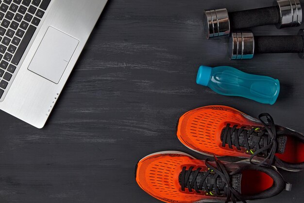 Orange sneakers dumbbells a water bottle and a laptop on a dark wooden background The concept of remote sports in quarantine view from above flat lay