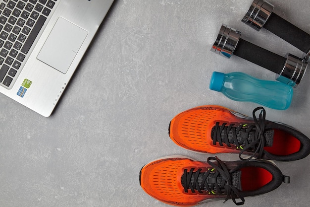 Orange sneakers dumbbells a water bottle and a laptop on a light gray concrete background Distance training in quarantine Top view with copy space Flat lay