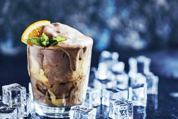 Orange Soda Creamsicle Ice Cream Float in glass decorated with piece of orange, straws and leaves on mint, surrounded by ice cubes on stone table