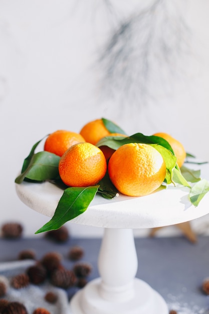 Оранжевые мандарины на сером фоне в новогоднем декоре с коричневыми сосновыми шишками и зелеными листьями. Новогоднее украшение с мандаринами. Вкусный сладкий клементин.