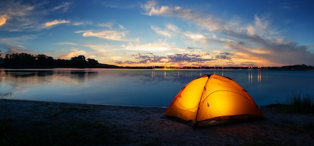 Tenda illuminata turistica arancione in riva al lago al tramonto