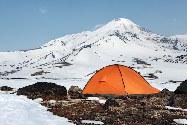 Orange tourist tent standing on meadow surrounded by snow on background of scenic volcanic cone