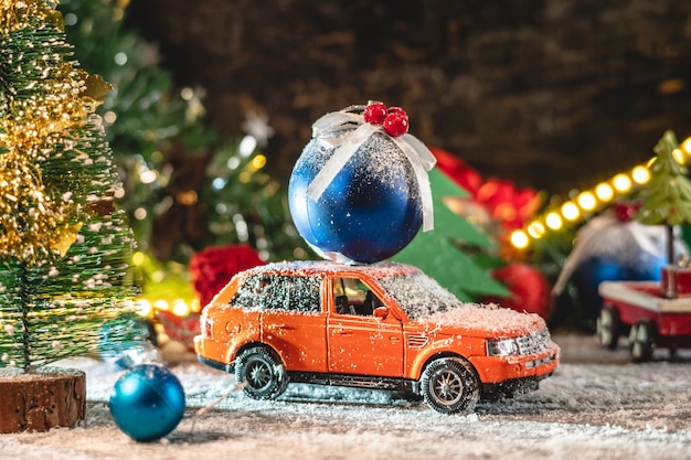 Orange toy car is going along a snow covered road and carrying a blue christmas ball