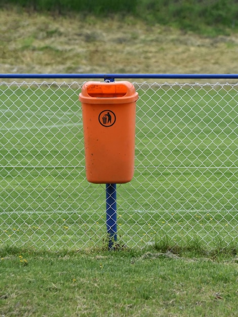 An orange trash can in the park attached to a metal wirexA fence