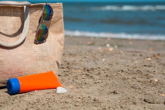 An orange tube with mock up sunscreen sunglasses and a beach bag stand on the sand near the sea and swing from the breeze UV protection beach holidays travel