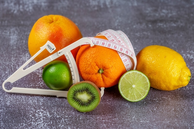 An orange wrapped with a measuring tape and a caliper surrounded by fresh fruits on a gray concrete background. The concept of slimming, remove cellulite, bring the figure into shape.