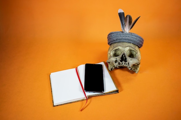 Oranje achtergrond met een open notitieboekje, een mobiele telefoon erop en een schedel met verendecoratie.