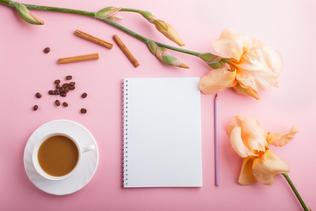 Oranje irisbloemen en een kop van koffie met notitieboekje op pastelkleurroze