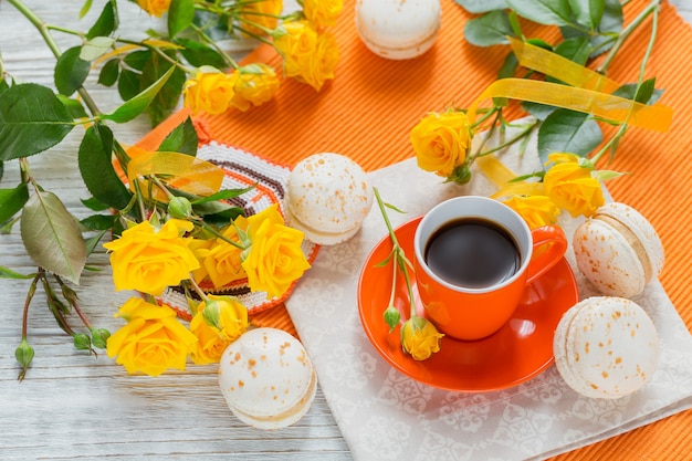 Oranje kopje zwarte koffie, gele rozen bloemen en zoete pastel Franse bitterkoekjes op witte houten tafel. Bovenaanzicht