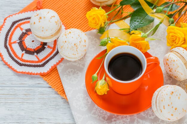 Oranje kopje zwarte koffie, gele rozen bloemen en zoete pastel franse bitterkoekjes op witte houten tafel