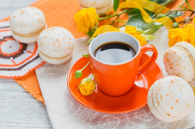 Oranje kopje zwarte koffie, gele rozen bloemen en zoete pastel franse bitterkoekjes op witte houten tafel