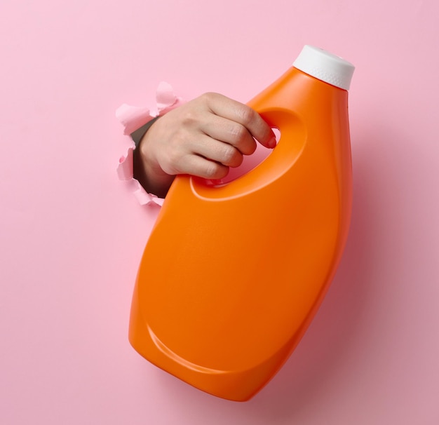 Oranje plastic fles met vloeibaar wasmiddel in een vrouwelijke hand op een roze achtergrond Een deel van het lichaam steekt uit een gescheurd gat op de achtergrond