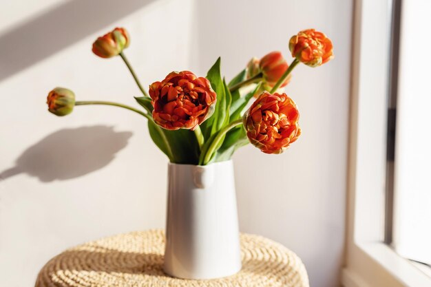 Oranje rode tulp bloemen met groene stengels en bladeren in een witte keramische kruik op een lichte schaduw witte muur en raam achtergrond Creatief bloemen plantkunde behang Minimale creatieve wenskaart