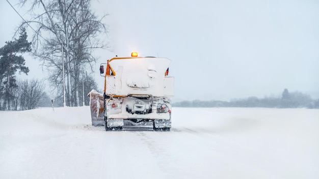 Oranje sneeuwploegwagen bedekt met sneeuw, op winterweg, grijze bewolkte hemelachtergrond, zicht van achteren