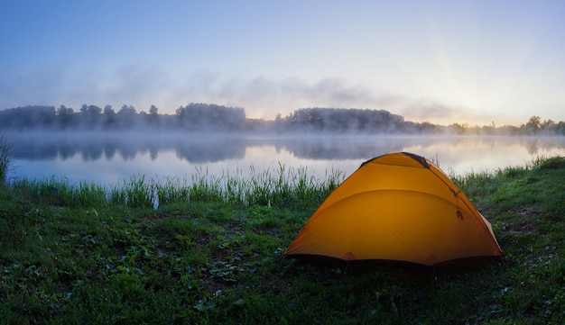 Oranje tent op groen gras van mistig meer bij zonsopgang