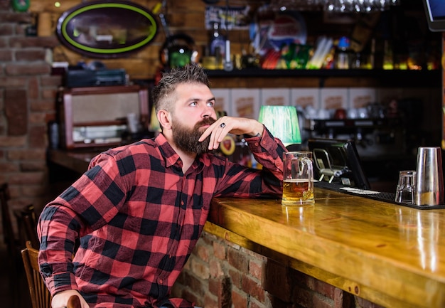 Order alcohol drink. Bar is relaxing place have drink and relax. Man with beard spend leisure in dark bar. Brutal hipster bearded man sit at bar counter drink beer. Hipster relaxing at bar with beer.