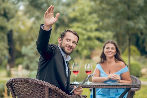 Order, cafe. Smiling young adult man in suit with menu raising hand and woman sitting in summer cafe