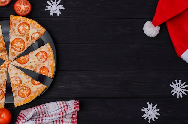 Ordering pizza for the Christmas day. Slices of pizza with cheese and tomatoes on a black plate with ingredients on a black background.