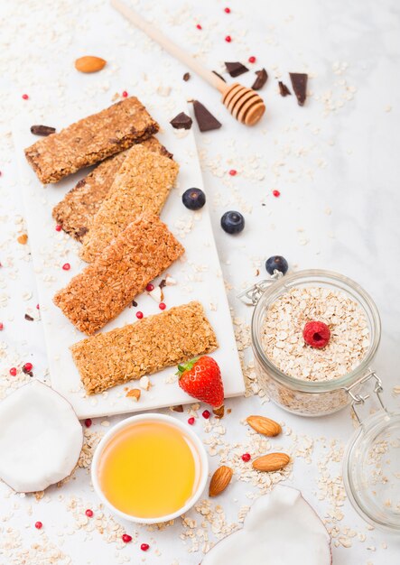 Organic cereal granola bar with berries on marble board with honey spoon and jar of oats and coconut on marble table.