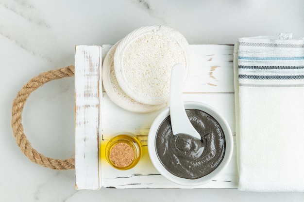 Organic cosmetic black clay essential oil and lufa csponges on a white textured tray The concept of spa and wellness Top view