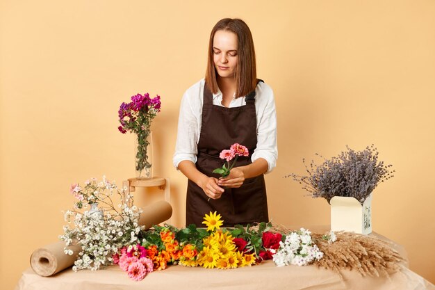 Organic flower market Aromatic blooms and plants Aromatic blooms and plants Brown haired woman florist working at her workplace wearing brown apron posing against beige wall