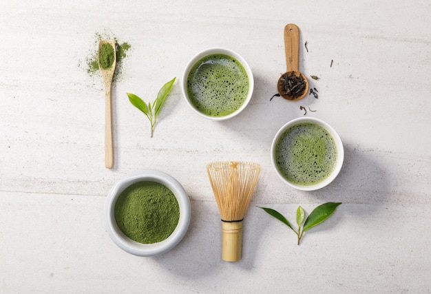 Photo organic matcha green tea powder in bowl with wire whisk and green tea leaf on white stone table