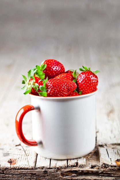 Organic red strawberries in white ceramic cup on rustic wooden background.