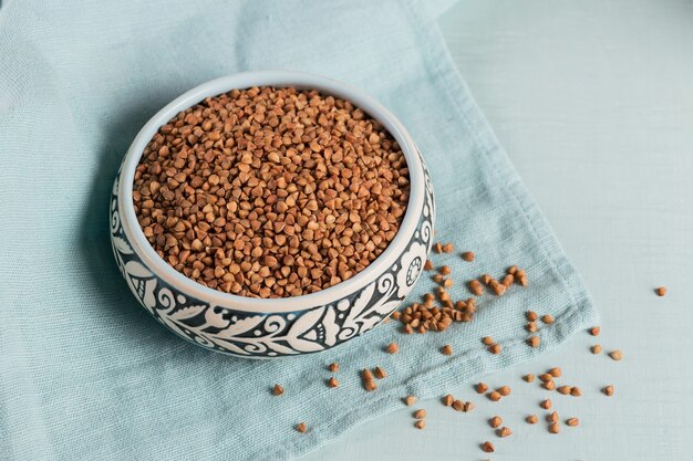 Organic uncooked scattered buckwheat grain in a bowl on a light blue background. Healthy and diet food concept.