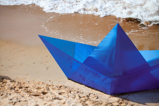 Origami blauwe papieren boot op zandstrand voor conceptontwerp, mooi papieren schip aan de wal. Modern kunstobject, close-up, oceaanachtergrond. Outdoor zomerplezier, reizen, zomervakantie concept