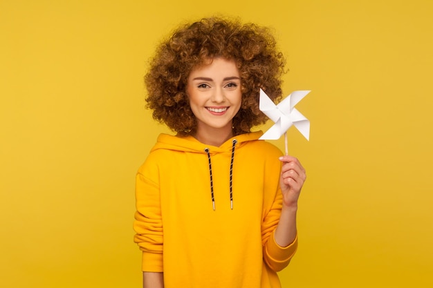 Origami handmolen. Portret van een gelukkige vrouw met krullend haar in een hoodie in stedelijke stijl die zorgeloos glimlacht en een papieren windmolen vasthoudt, een pinwheel-speelgoed op een stok. indoor studio-opname geïsoleerd op gele achtergrond