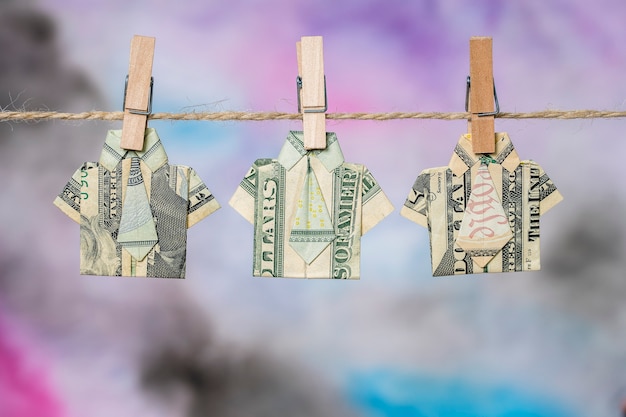 Origami shirt gemaakt van dollar biljet op hangende kleurrijke achtergrond. Dollarbiljetten hangen aan een touw. Detailopname. T-shirt met dollarbiljet