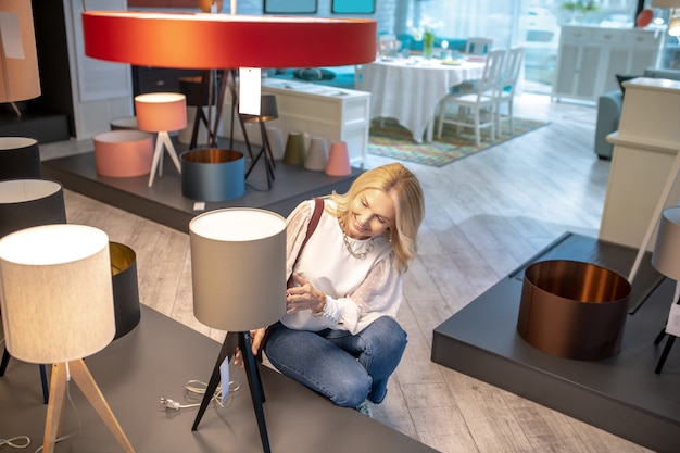 Original floor lamp. Good looking woman crouched near a beige floor lamp, looking at him, in a delightful mood