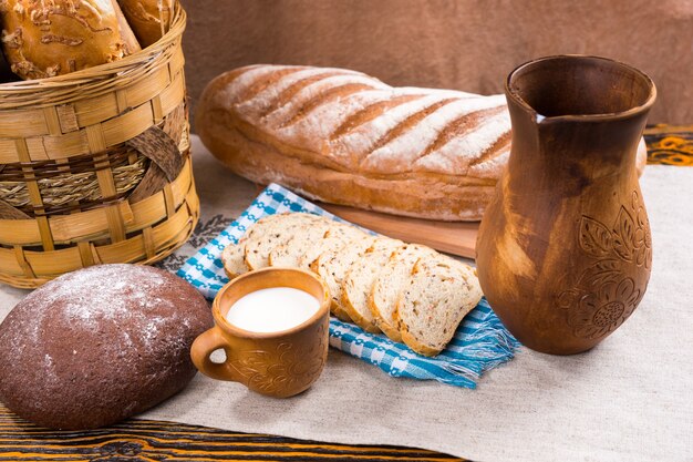 Ornately decorated wooden pitcher, little cup of milk and freshly baked bread as whole loaf and slices next to basket on table